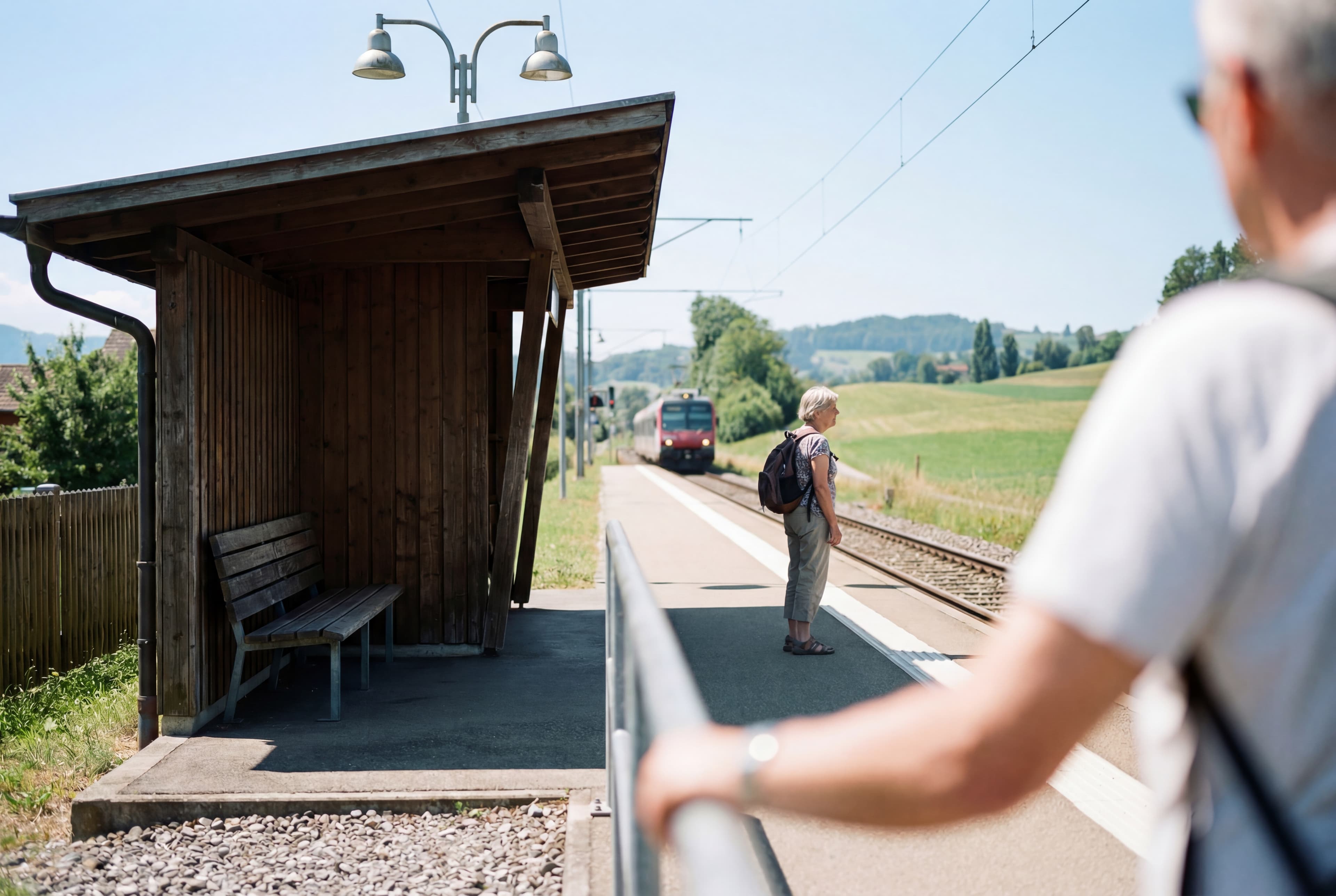 Older woman with a backpack standing on a rural train platform, looking toward an approaching train, with green fields and overhead railway lines in the background.
