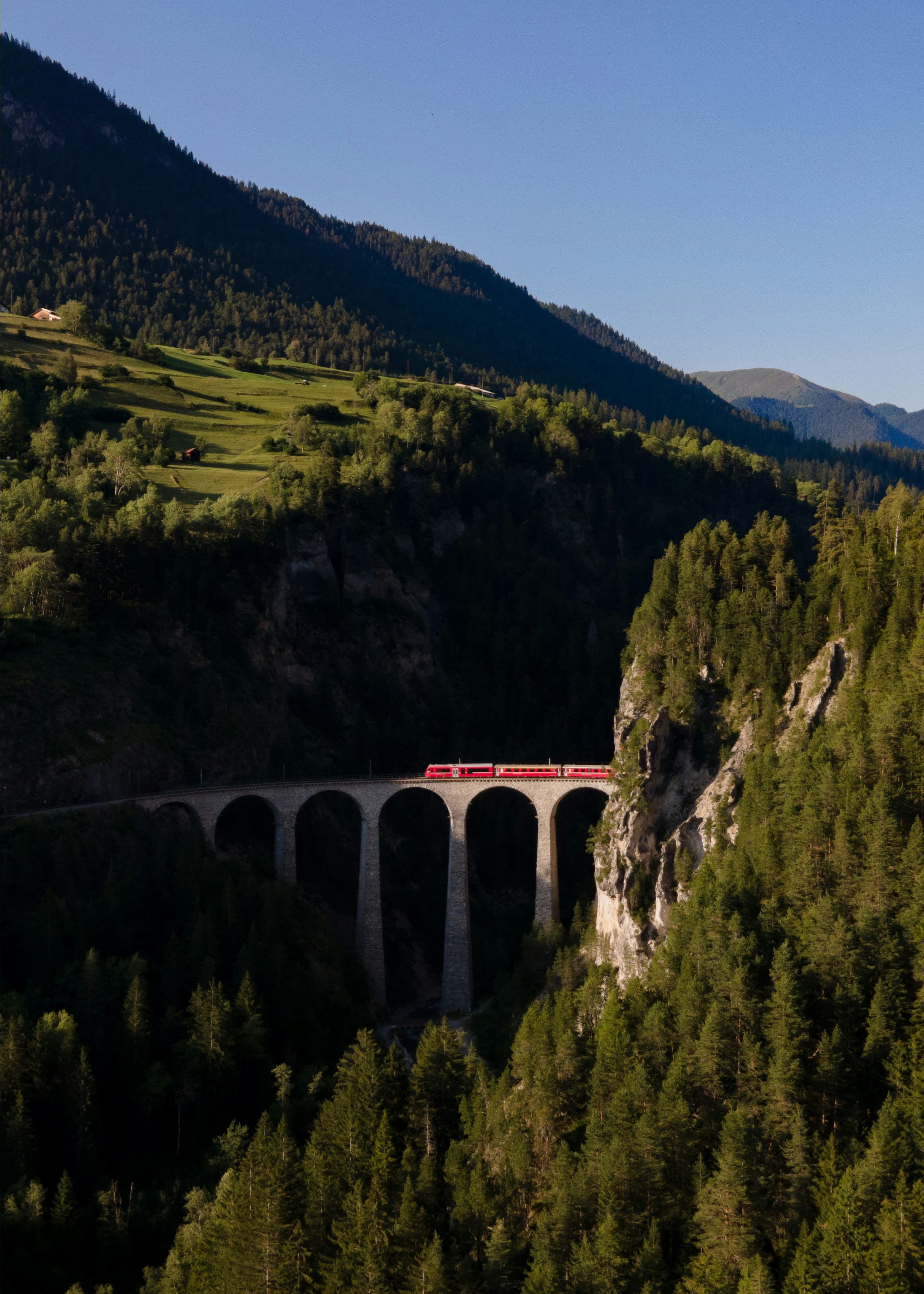 A red train crossing a tall stone viaduct surrounded by dense forest and steep mountain slopes.